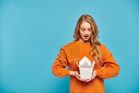 A beautiful blonde woman in an orange sweater joyfully holding a box of delicious Asian food.