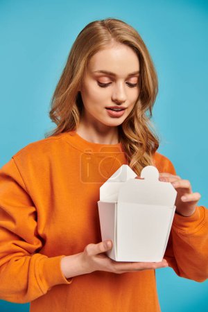 Foto de Una hermosa mujer en un suéter naranja sostiene una caja de comida blanca, su expresión curiosa y encantada. - Imagen libre de derechos