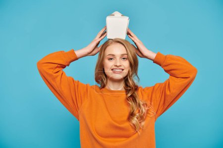Photo for A beautiful woman in an orange sweater joyfully holds box of Asian food over her head. - Royalty Free Image