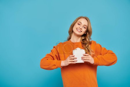 Eine stilvolle Frau in orangefarbenem Pullover genießt einen Moment der Entspannung, während sie eine Lebensmittelbox in der Hand hält.