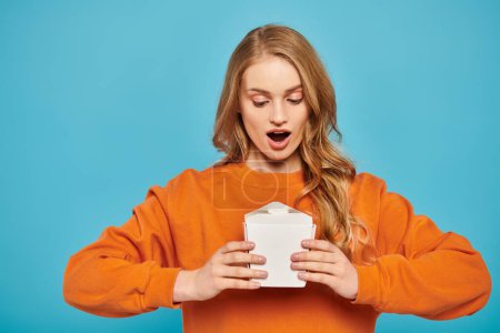 A stunning blonde woman in an orange sweater holds a white food box