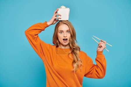 A beautiful blonde woman in an orange sweater playfully holds chopsticks over her head with a food box nearby.