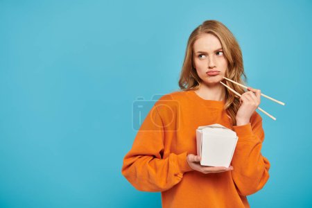 Photo for Blonde-haired woman gracefully holds chopsticks and a box of delicious Asian food. - Royalty Free Image