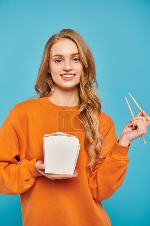 A beautiful woman with blonde hair delicately holds food box and chopsticks, savoring Asian cuisine.