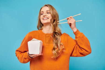 A blonde woman gracefully holds chopsticks and a box of Asian food, showcasing a blend of beauty and culinary artistry.