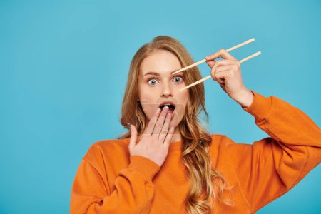 A beautiful blonde woman holding two chopsticks over her mouth, ready to enjoy Asian cuisine.