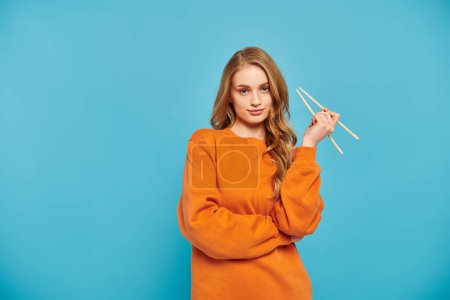 A beautiful woman in an orange sweater delicately holds a pair of chopsticks, ready to enjoy a delicious Asian meal.
