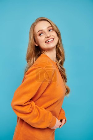Photo for A graceful woman in an orange sweater posing on blue vibrant backdrop. - Royalty Free Image