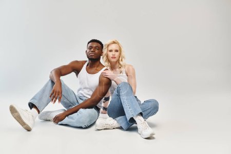 A young multicultural couple sitting on the ground in a studio, embodying peaceful togetherness amidst a grey backdrop.