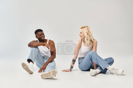 Ein junges multikulturelles Paar sitzt friedlich und verbunden auf dem Boden in einem Studio vor grauem Hintergrund.