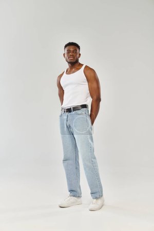 A young and sexy African American man confidently poses in a white tank top and jeans against a grey studio backdrop.