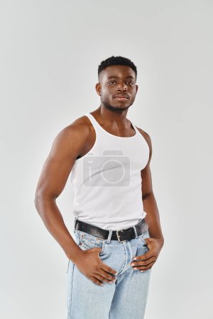 Téléchargez les photos : Un jeune Afro-Américain sexy en chemise blanche et en jean pose dans un studio sur un fond gris. - en image libre de droit