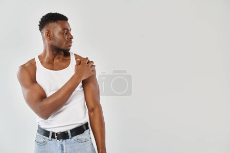 A young, sexy African American man in a white tank top and jeans poses confidently in a studio against a grey background.