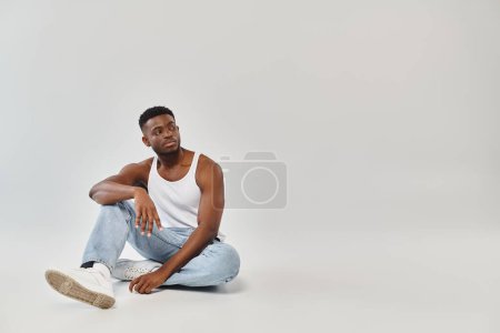 Junger Afroamerikaner sitzt mit gekreuzten Beinen im Studio vor grauem Hintergrund.