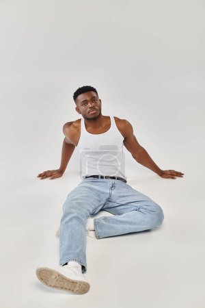 Ein junger und sexy afroamerikanischer Mann sitzt mit ausgestreckten Armen auf dem Boden in einem Studio vor grauem Hintergrund.