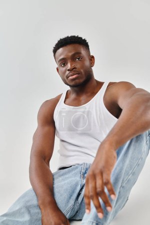 A young African American man in a white tank top sits on the ground in a contemplative pose, against a neutral grey background.