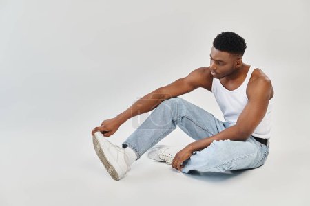 Ein junger, stylischer afroamerikanischer Mann sitzt in lässiger Jeans und Tank-Top ruhig auf dem Boden.