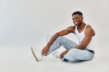 Young African American man sitting on the ground, legs crossed, in a studio setting against a grey background.