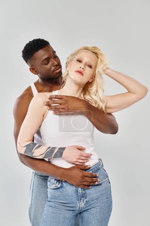 Ein Mann hält eine Frau zärtlich auf dem Arm und drückt damit Liebe und Intimität aus. Sie sind ein junges interrassisches Paar auf grauem Studiohintergrund.