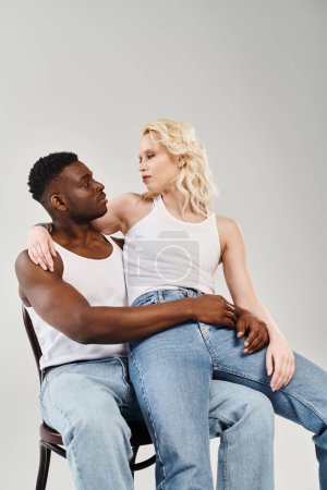 A young interracial couple leisurely seated on a chair in a studio against a grey background.