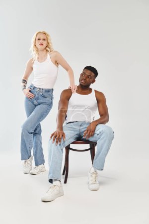 Téléchargez les photos : Un jeune couple interracial assis sur une chaise dans un studio sur fond gris, dégage un sentiment d'unité et de convivialité. - en image libre de droit