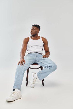 Photo for A young man in a white tank top sitting on a chair in a studio with a grey background. - Royalty Free Image
