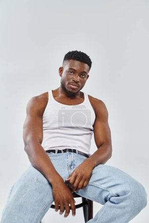 A young man of an interracial couple sits confidently atop a chair while wearing jeans on a grey studio background.