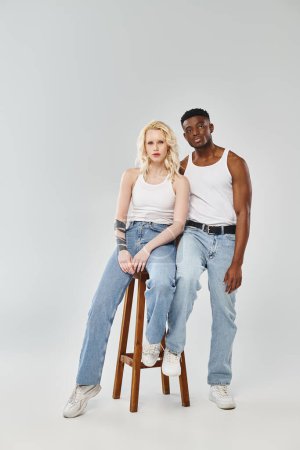 A young interracial couple sits gracefully together on a stool in a studio against a grey background.