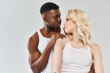 Téléchargez les photos : Jeune couple interracial, homme et femme, debout ensemble dans des débardeurs blancs dans un studio sur fond gris. - en image libre de droit