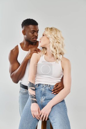 A young interracial couple standing shoulder to shoulder in a studio against a grey background.