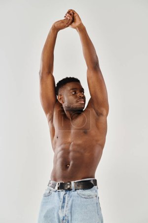 A young, shirtless African American man joyfully raises his hands in surrender on a grey studio background.