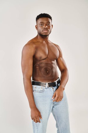 Un joven afroamericano sin camisa se encuentra confiado en jeans, mostrando su físico tonificado sobre un fondo gris.