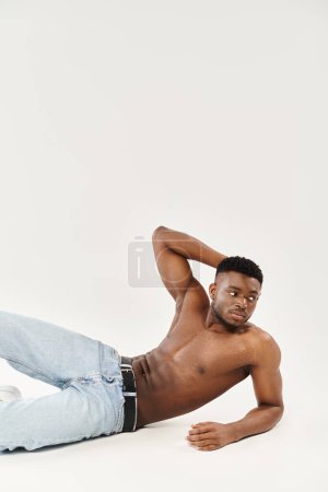 Un joven afroamericano yace tendido sin camisa en el suelo, exudando sensualidad y tranquilidad en un ambiente de estudio.