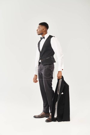 Young African American groom confidently holding a jacket, styled in a sleek suit and tie against a grey studio backdrop.