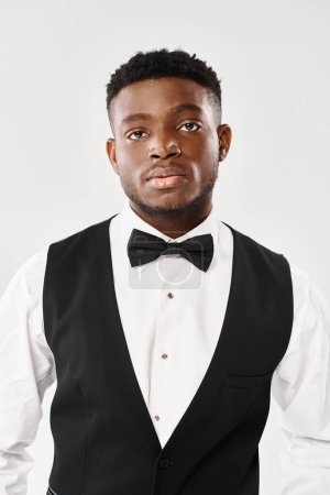 Young African American groom exudes style in a black vest and bow tie against a grey studio background.