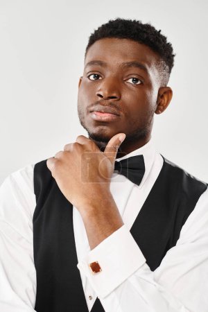 Handsome young African American groom wearing a black vest and bow tie in a studio setting against a grey background.