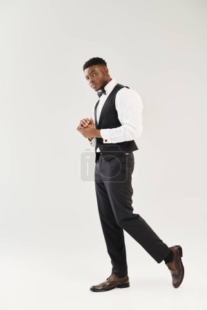 Ein junger, gutaussehender afroamerikanischer Bräutigam in Anzug, Weste und Krawatte posiert in einem Studio vor grauem Hintergrund.