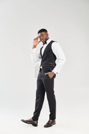 Un joven y guapo novio afroamericano vestido con un chaleco y corbata negros, exudando elegancia y encanto en un estudio rodado sobre un fondo gris.