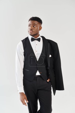 Un joven y guapo novio afroamericano toma una pose confiada en un elegante esmoquin contra un fondo gris de estudio.