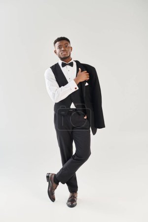 A young, handsome African American groom in a tuxedo poses stylishly in a studio against a grey background.