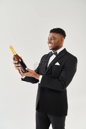 Schöner afroamerikanischer Bräutigam im eleganten Smoking, hält eine Flasche Champagner in der Hand und strahlt Eleganz im Studio-Ambiente aus.