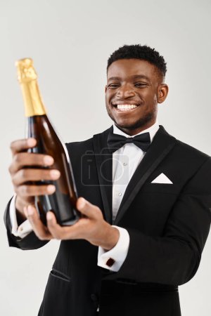 Foto de El novio afroamericano en esmoquin disfruta de una botella de champán en un estudio sobre un fondo gris. - Imagen libre de derechos