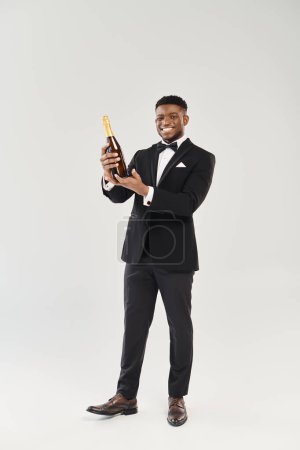 Guapo novio afroamericano en esmoquin sostiene botella de champán, preparado para brindis de celebración.