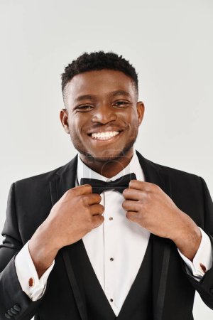 Handsome African American groom in tuxedo smiling as he adjusts his bow tie against a grey studio background.