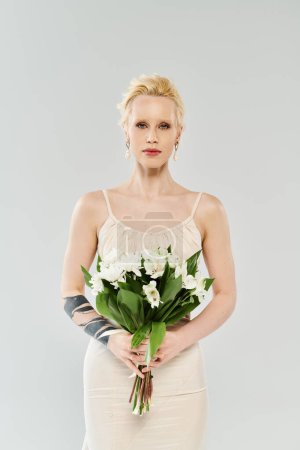 Ätherische blonde Braut in weißem Kleid, anmutig in der Hand einen lebendigen Blumenstrauß vor grauem Hintergrund.