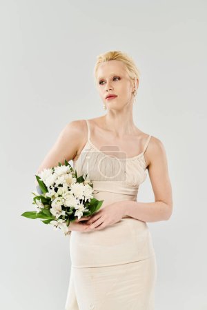 Une belle mariée blonde en robe tient un bouquet de fleurs dans un studio sur fond gris.