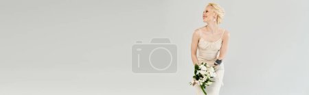 A stunning blonde bride stands gracefully in a white dress, delicately holding a lush bouquet of vibrant flowers.