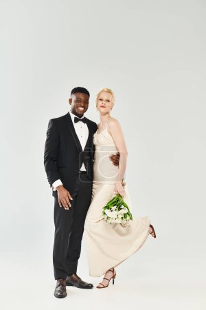 Eine schöne blonde Braut im Hochzeitskleid und ein afroamerikanischer Bräutigam, beide in formeller Kleidung, posieren elegant für ein Porträt in einem Studio vor grauem Hintergrund.