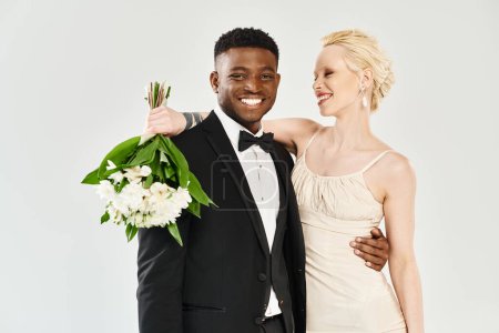 A beautiful blonde bride in a wedding dress and an African American groom in a tuxedo standing confidently together in a studio.