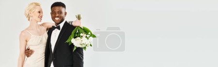 African American groom in tuxedo stands proudly next to beautiful blonde bride in white dress, exuding grace and style.